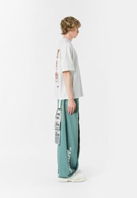 Camiseta blanca oversized con estampado gráfico en la parte posterior, combinada con pantalones sueltos en tono verde azulado que presentan una banda lateral de contraste y una etiqueta en el muslo.