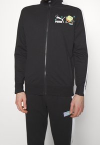 Veste de survêtement noire Puma avec fermeture éclair à l'avant, bandes blanches sur les manches et motif SpongeBob SquarePants sur le côté gauche de la poitrine.