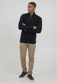 Junger Mann mit verschränkten Armen, der einen schwarzen Reißverschluss-Pullover, beige Cargo-Hosen und schwarze Sneakers trägt und vor einem schlichten Hintergrund steht.