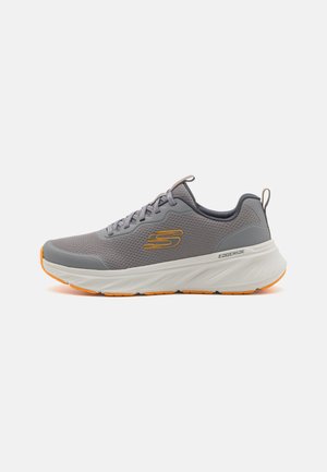 Shoe atlética cinza com parte superior de malha, detalhes laranja e sola branca texturizada. Apresenta bico arredondado e design com atacadores.