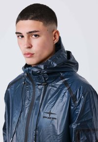 Giovane uomo con capelli corti che indossa una giacca impermeabile lucida di colore blu scuro con cappuccio e logo PXP, rivolto in avanti con un'espressione neutra.