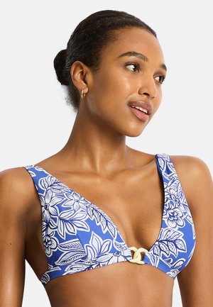 Donna con capelli scuri raccolti in uno chignon, che indossa orecchini a cerchio dorati e un top bikini blu con motivo floreale e chiusura color crema, guarda verso destra.