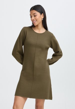 Vrouw met lang donker haar draagt een olijfgroene, gebreide jurk met lange mouwen, staat en lacht tegen een effen lichte achtergrond.