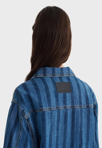 Personne aux cheveux longs et bruns vue de dos, portant une veste en denim rayée bleue avec une étiquette noire rectangulaire sur le haut du dos.