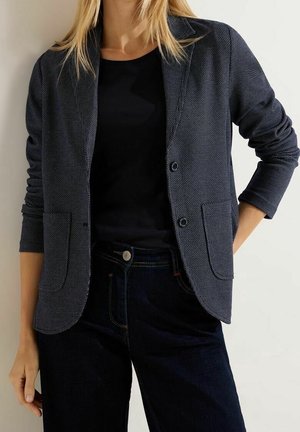 Frau trägt strukturierten dunkelgrauen Blazer über schwarzem Hemd und dunklen Jeans, steht vor einfachem hellem Hintergrund.