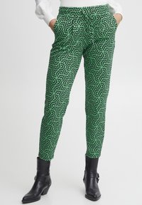 Grüne, gemusterte Hose mit einem geometrischen schwarzen Design, schmaler Schnitt, elastischem Bund und Seitentaschen, kombiniert mit schwarzen Knöchelstiefeln.