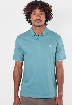 Homme aux cheveux bouclés portant un polo bleu clair à manches courtes avec un petit logo blanc sur la poitrine gauche sur fond blanc.
