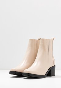 Bottines en cuir beige avec une finition lisse, bout rond, talon structuré et détails élastiques sur le côté. Semelles en caoutchouc noir.