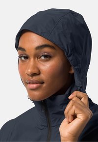 Marineblauwe waterdichte jas met een hoge kraag en een verstelbare capuchon. Soepele textuur en een rits aan de voorkant. Model houdt de kraag vast en toont de pasvorm.