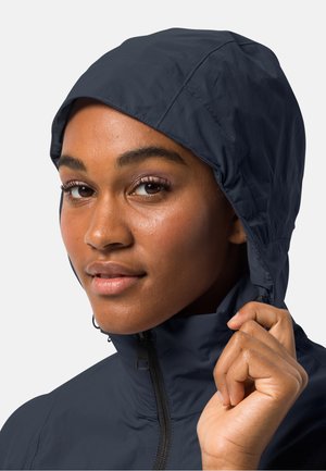 Veste imperméable bleu marine avec un col haut et une capuche réglable. Texture lisse et zip frontal. Le modèle tient le col, mettant en valeur l'ajustement.