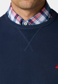 Marineblauwe sweatshirt met een gestructureerde brei en kruissteekdetails bij de halslijn, laag over een geruit overhemd met blauw, rood en witte patronen.