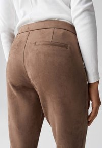 Pantaloni in suede marrone con un design elegante, dotati di una tasca posteriore e una texture liscia, abbinati a una maglietta bianca a maniche lunghe.