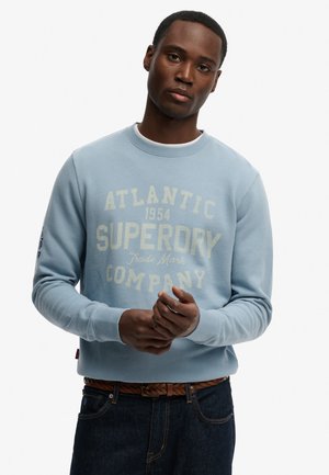 Giovane uomo che indossa una felpa azzurra con la scritta "Atlantic 1954 Superdry Company", in piedi con le mani giunte davanti a uno sfondo bianco.