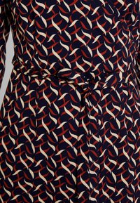 Marinblå klänning med ett repetitivt mönster av vita och röda kurviga linjer, med ett knytbälte i midjan och mjuktexturerad tyg.