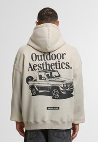 Uomo che indossa una felpa beige con il testo "Outdoor Aesthetics" e un'illustrazione nera di un veicolo fuoristrada sulla schiena.