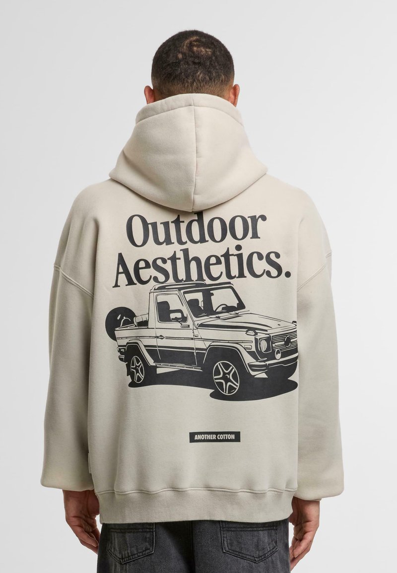 Uomo che indossa una felpa beige con il testo "Outdoor Aesthetics" e un'illustrazione nera di un veicolo fuoristrada sulla schiena.