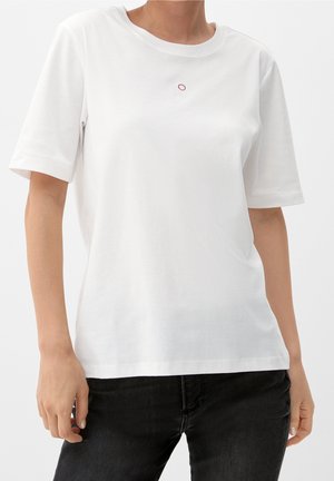 Kvinna som bär en enkel vit T-shirt med kort ärm och en liten röd och vit broderad design vid halsringningen, i kombination med svarta jeans.