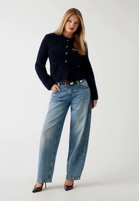 Marineblaue, strukturierte Jacke mit Knöpfen, kombiniert mit hellblauen, weitem Jeans. Das Modell trägt dunkle High Heels und einen schwarzen Gürtel mit silberner Schließe.