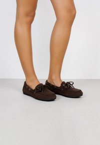 Zapatos de ante marrón en estilo mocasín con puntera redondeada, detalle de cordones y ojales de plata. La suela es plana y flexible.