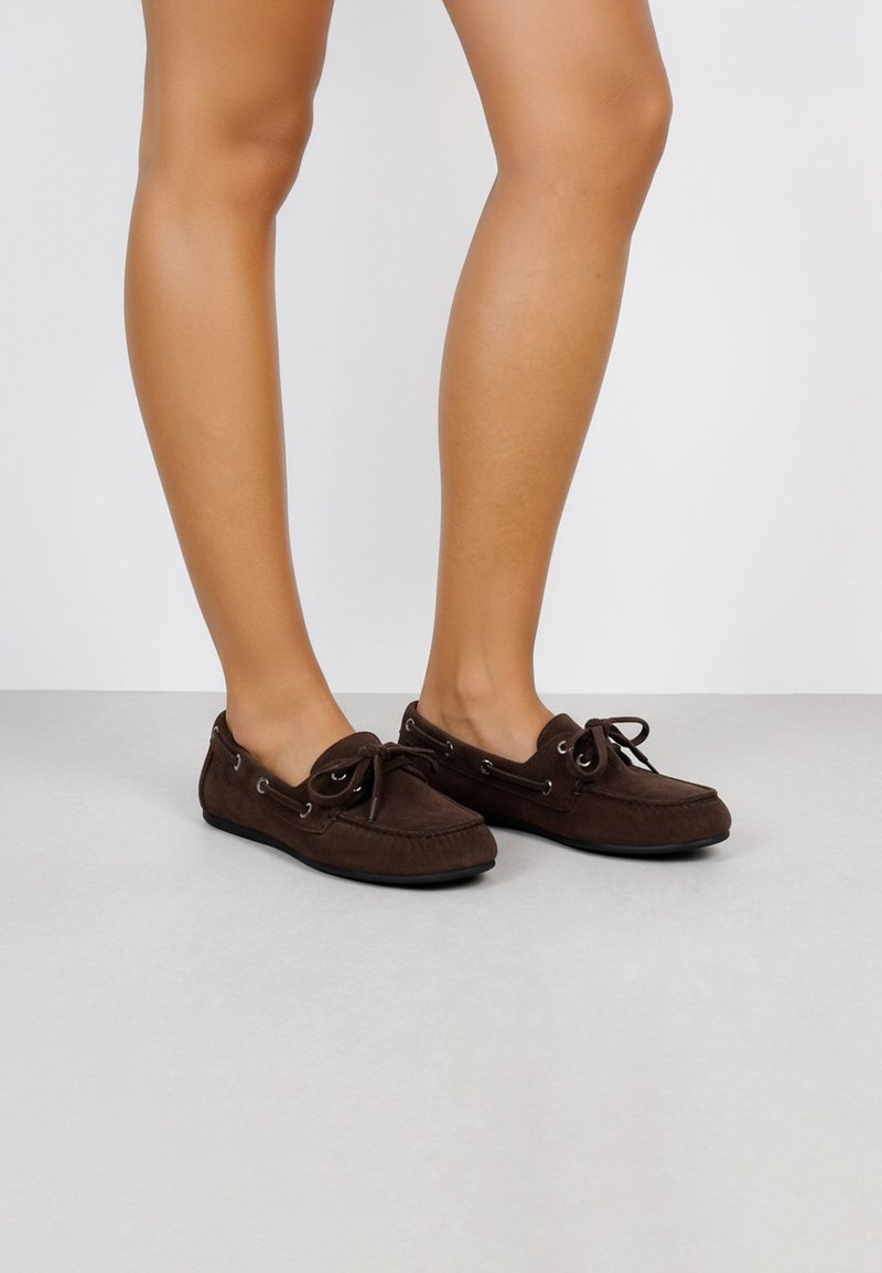 Zapatos de ante marrón en estilo mocasín con puntera redondeada, detalle de cordones y ojales de plata. La suela es plana y flexible.