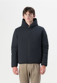 Schwarze isolierte Jacke mit hohem Kragen und Reißverschlusstasche auf der Brust, mit glatter Textur und tailliertem Design. Getragen über hellen Hosen.