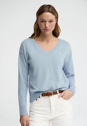 Kobieta o jasnobrązowych włosach, ubrana w jasnoniebieski sweter z dekoltem w serek, białe spodnie i brązowy pasek, stojąca z jedną ręką w kieszeni.