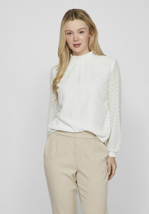 Woman with long blonde hair wearing a white patterned long-sleeve blouse and beige high-waisted pants, standing against a plain background.