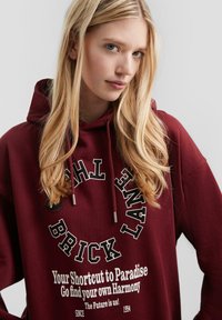 Giovane donna con lunghi capelli biondi che indossa un hoodie bordeaux con testo nero e bianco che recita "BRICK LANE" e altre frasi motivazionali.