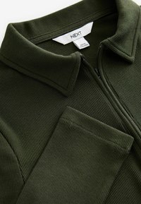 Oliivinvihreä, ribattu vetoketjupaita, jossa on käännettävä kaulus ja pitkät hihat. Varustettu vetoketjulla ja tarralla, jossa on teksti "NEXT."