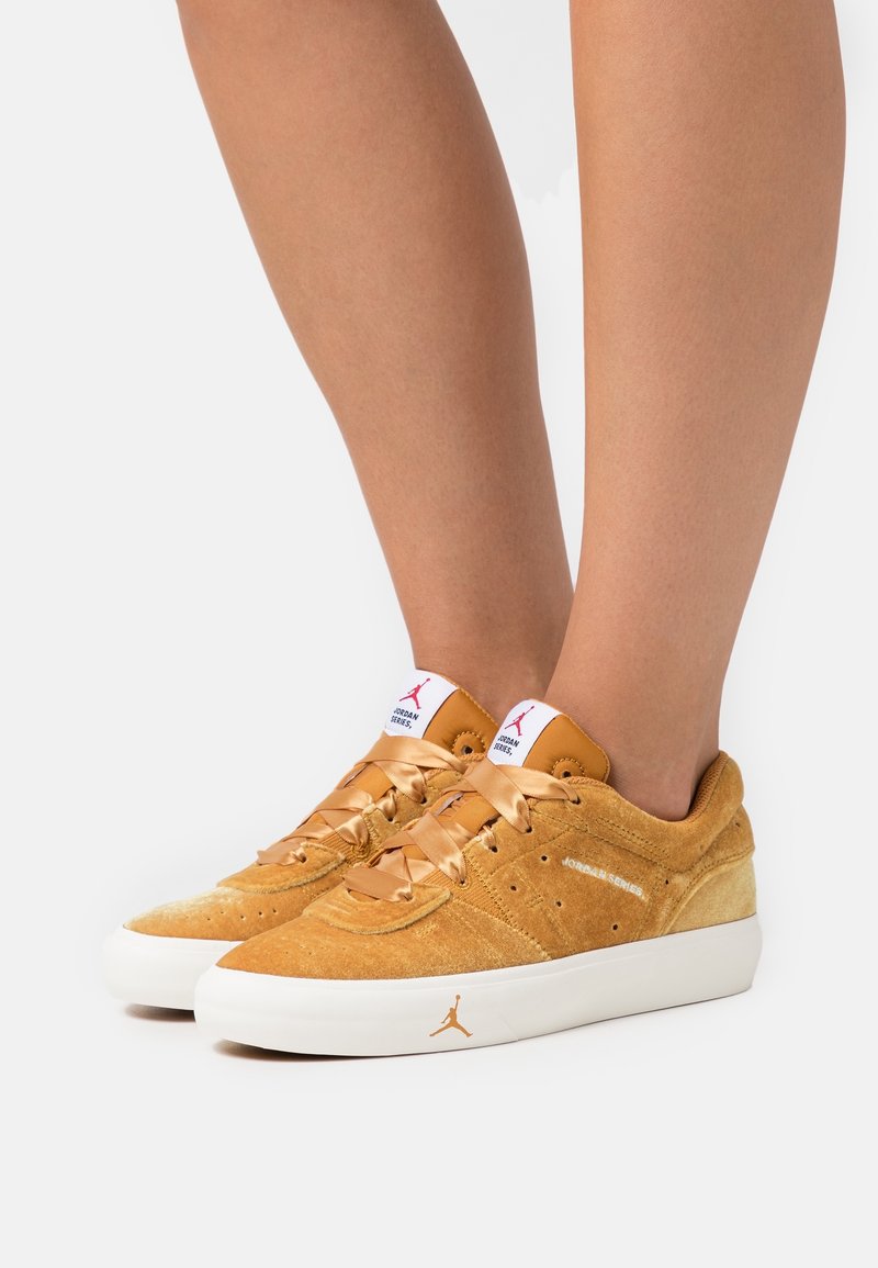 Tan mockasnören med låg profil, som har texturerade detaljer, satin snören och en vit gummisula med en logotyp på sidan.