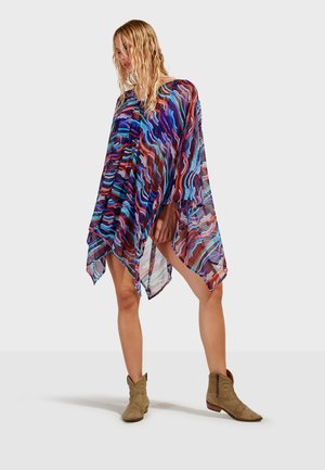 Donna che indossa un abito poncho trasparente multicolore con motivi e stivaletti in camoscio nocciola, in piedi davanti a uno sfondo semplice.