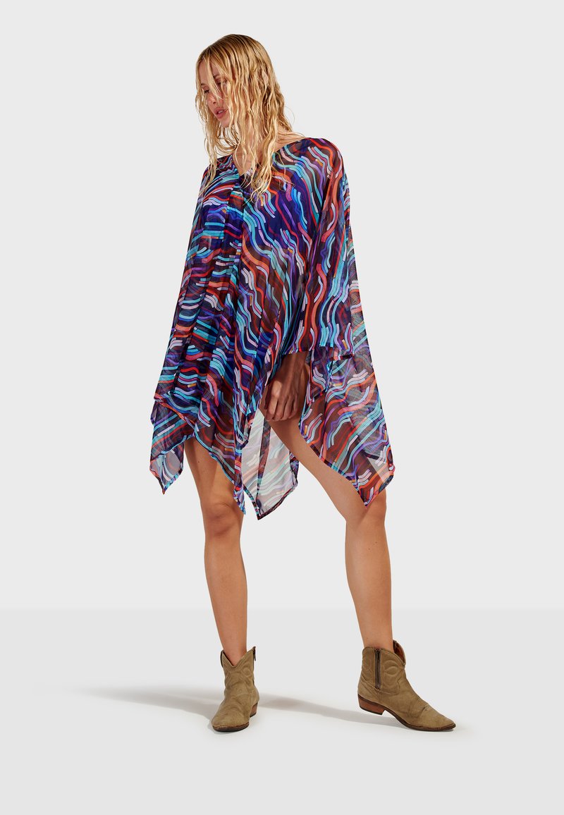 Femme portant une robe poncho transparente multicolore à motifs et des bottines en daim beige, debout devant un fond uni.