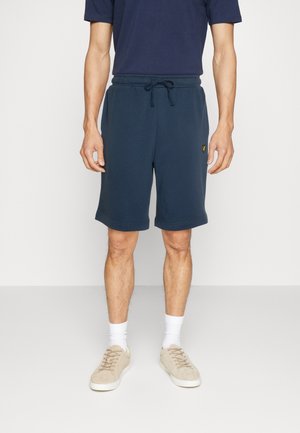 Man draagt marineblauwe shorts met trekkoord, marineblauw T-shirt, witte sokken en beige sneakers, staand op een effen lichte achtergrond.