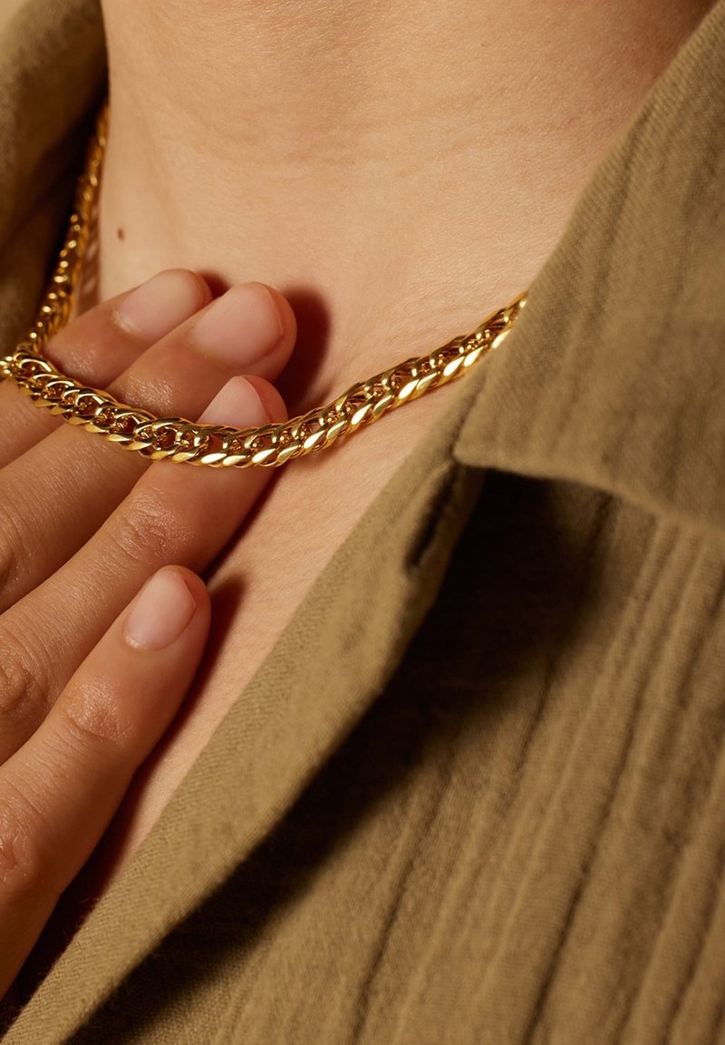 Gouden ketting met gedraaide schakels, rustend op de huid, handen die het aanraken. Op de achtergrond is een textuur van een olijfgroen kledingstuk te zien.