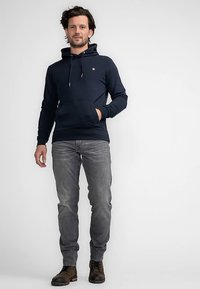 Sudadera con capucha azul marino con cordones y bolsillo delantero, combinada con vaqueros grises y zapatos marrones. La tela parece suave y lisa. Incluye un logo.