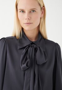 Mörkgrå blus med en stor sljust vid kragen, med puffärmar och en mjuk, slät tygkänsla.