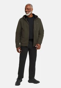 Dunkelgrüne Fleecejacke mit Reißverschluss vorne und Kapuze, kombiniert mit schwarzer Hose und schwarzen Schuhen. Das Modell trägt eine lockere Passform.