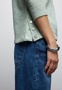 Jasnozielony sweter o teksturze z trzema metalowymi guzikami po boku, zestawiony z ciemnoniebieskimi dżinsami. Proste, codzienne elementy designu.