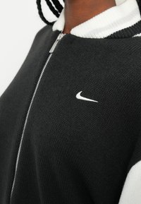 Črna zip-up jakna z belim rebrastim ovratnikom in manšetami. Ima teksturirano tkanino in majhen bel Nike logotip na prsih.