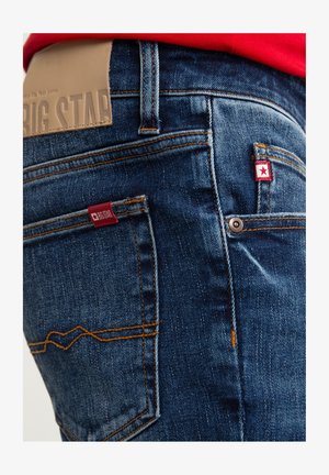 Niebieskie dżinsy z denimu o fakturowanym wykończeniu, z jasnobeżową skórzaną łatą i czerwoną metką z logo na tylnej kieszeni. Kontrastowe szwy.