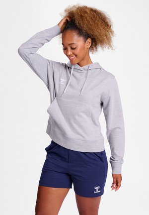 Kvinde med krøllet hår iført en lysegrå hættetrøje og marineblå shorts, smilende og poserer med den ene hånd på hovedet.