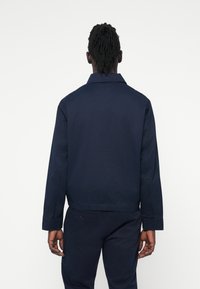 Marineblå jakke med lige snit, knaplukning ved ærmerne og klassisk krave. Lavet af robust stof, glat tekstur, minimalistisk design.