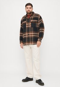 Flannel shirt in black with brown and orange plaid pattern, featuring chest pockets, button closure, and relaxed fit; paired with cream pants and black shoes.