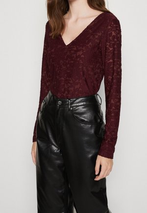 Femme portant un haut en dentelle bordeaux ajusté à manches longues avec des motifs floraux et un pantalon noir en similicuir taille haute.