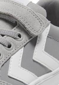 Sneaker grigio realizzato in materiali scamosciati e pelle liscia, con strisce bianche, lacci rotondi e una chiusura con strap in velcro.