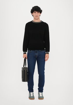 Sort sweater med kontrastkrave, blå jeans, grønne og hvide sneakers, holder en sort cylindrisk taske med rem. Neutral baggrund.