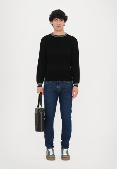 Schwarzer Pullover mit kontrastierendem Kragen, blaue Jeans, grüne und weiße Turnschuhe, hält eine schwarze zylindrische Tasche mit einem Riemen. Neutraler Hintergrund.