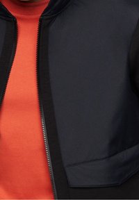 Giacca a zip blu navy con una texture di tessuto morbido e una felpa rossa in contrasto sotto; presenta un colletto rialzato e una tasca laterale.