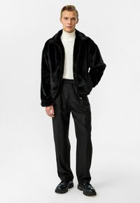 Zwarte nepbont jas met ritssluiting, gedragen over een witte coltrui. Gepaard met zwarte nette broek en zwarte schoenen.