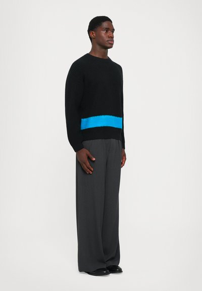 Schwarzer gestrickter Pullover mit einem breiten, leuchtend blauen horizontalen Streifen auf der Taille. Kombiniert mit lockeren, grauen Hosen und schwarzen Schuhen.